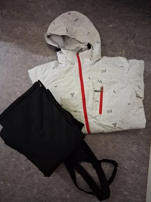Skidjacka och byxor. Storlek XS - Säljer ett set med en vit skidjacka från med grått geometriskt mönster och röda dragkedjor. Jackan har huva med snörning. Svarta termobyxor med fickor och dragkedja nere vid fotändan.