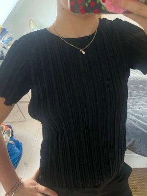 Svart ribbad topp med glittereffekt - Säljer en svart topp med korta ärmar och rund hals. Tyget är ribbat och har en glittrig finish som ger en snygg effekt. Passformen är normal och materialet känns mjukt och stretchigt, perfekt till många olika looks.