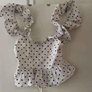 Vit prickig topp med volangärmar från H&M - Supersöt vit topp med svarta prickar från H&M. Toppen har stora volangärmar och en volang nertill samt knytning i ryggen. Perfekt för dig som gillar lekfulla detaljer och trendig stil.