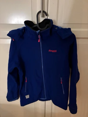 Blå vindjacka från Bergans - Blå vindjacka från Bergans med huva och snygga röda detaljer på dragkedjor och broderad logga. Jackan har två fickor med dragkedja och är tillverkad i slitstark polyester. Köp nu jac postar snabbt!!💕💗