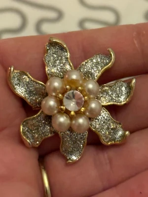 Glittrig blomformad brosch med pärlor - Säljer en unik brosch i form av en blomma med glittriga silverblad och guldfärgade kanter. I mitten sitter skimrande pärlor och en stor kristall. Perfekt som statement-accessoar för att lyfta din outfit.