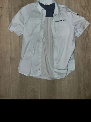 Ljusblå kortärmad skjorta från H&M - Säljer en ljusblå skjorta från H&M i storlek M. Skjortan har korta ärmar, två bröstfickor med knappar och trycket 'SUPERIOR NEW GOODS' på ena fickan. Tillverkad i mjuk bomull och har klassisk krage med kontrastfärgad insida.