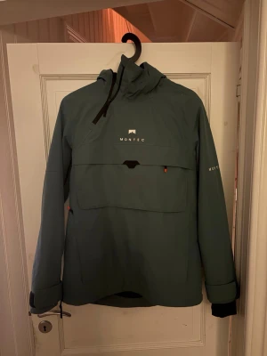 Skidjacka från Montec - Snygg grön vindjacka från Montec med stor ficka framtill, dragkedja i sidan och justerbar huva. Jackan har långa ärmar med muddar och coola detaljer som logga på bröstet och ryggen. Perfekt för dig som gillar skidåkning! Jackan är endast använd en säsong med fåtal åk. ☺️