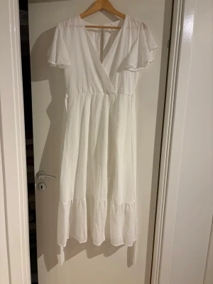 Vit långklänning med volang - Säljer en vit långklänning i storlek S med omlott-v-ringning och korta, luftiga ärmar. Klänningen har volangdetaljer nedtill och är tillverkad i ett lätt material som ger ett flowigt intryck. Perfekt för sommarens alla tillfällen.