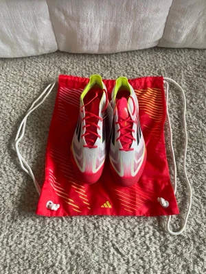 Adidas F50 ELITE FG fotbollsskor Storlek 42 - Säljer ett par Adidas X Speedflow F50 fotbollsskor i en riktigt fräsch färgkombo med vit, röd och neon-gul. Skorna har snörning, platt sula med dobbar och är tillverkade i syntetmaterial. Perfekta för dig som vill ha grym känsla och speed på planen. Den kommer även med en påse. Ordinarie pris:3099kr
