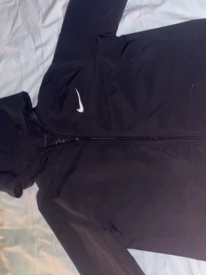 Svart Nike vindjacka  - Svart vindjacka från Nike i storlek XL (158-170) med huva och hel dragkedja framtill. Jackan har en stilren design med vit Nike-logga på bröstet och är tillverkad i lätt polyester med Dri-Fit-teknologi som håller dig torr. Perfekt för aktiva dagar och sport.