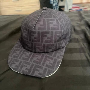 Svart Fendi keps med logomönster - Snygg svart keps från Fendi med deras ikoniska FF-logga över hela kepsen. Klassisk basebollmodell i 100% polyester med böjd skärm och justerbar baksida. Perfekt för dig som vill ha en stilren och lyxig accessoar med streetkänsla.