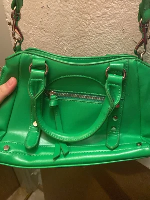 Grön axelväska med dragkedja - Cool grön axelväska i syntetmaterial med både handtag och justerbar axelrem. Väskan har en snygg silvrig dragkedja framtill och detaljer i metall. Perfekt storlek för det viktigaste och ger en färgklick till din outfit.