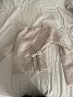 Beige omlott kofta med knyt - Mysig beige omlott kofta från NA-KD med v-ringning och knyt i midjan. Koftan är stickad med ribbade detaljer vid ärmslut och nederkant, och har långa ärmar. Perfekt för lager-på-lager under kyliga dagar.