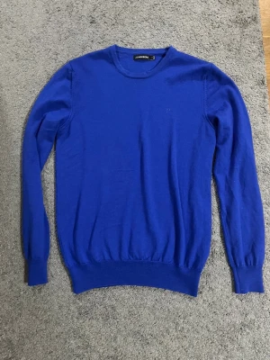  J.Lindeberg tröja - Snygg blå stickad tröja från J.Lindeberg med rund hals och diskret logga på bröstet. Tröjan har långärmade ribbade muddar och är perfekt för dig som gillar stilrena plagg med enkel design.