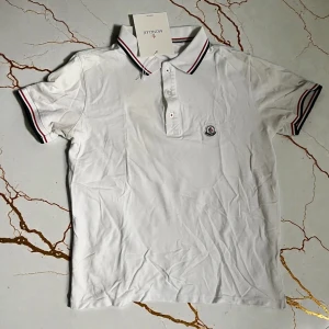 Vit Moncler piké med rödblå detaljer - Säljer en vit pikétröja från Moncler med klassisk krage och knappslå framtill. Pikén har röda och blå ränder på krage och ärmslut samt Moncler-logga på bröstet. Materialet är mjuk bomull och modellen är kortärmad med normal passform.