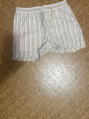 Linneshorts - Säljer ett par randiga shorts i vitt och beige med snörning i midjan. De har en relaxed passform, fickor fram och en bakficka. Perfekta för varma dagar och chill häng. Materialet känns lätt och skönt mot huden.