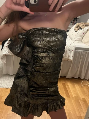 Glittrig guld kortklänning med volang - Säljer en glittrig guld kortklänning med snygga volangdetaljer på sidan och längs nederkanten. iguren. Perfekt nu till nyår ❤️❤️(aldrig använd )den är i storlek m men passar absolut s med !!