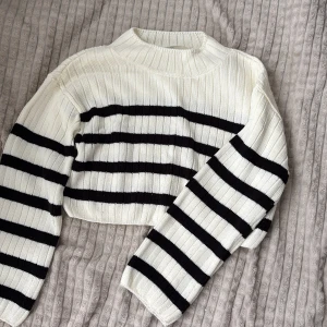 Randig croppad stickad tröja H&M - Säljer en croppad stickad tröja från H&M i vitt med svarta horisontella ränder. Tröjan har ribbad struktur, lång ärm och rund halsringning. Perfekt för lager-på-lager och chill stil.