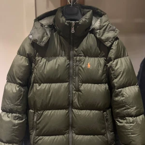 Polo Ralph Lauren pufferjacka olivgrön - Olivgrön pufferjacka från Polo Ralph Lauren med orange broderad logga på bröstet. Jackan har huva, dragkedja framtill och två sidofickor med dragkedja. Fylld för extra värme och perfekt för kyliga dagar. Klassisk och stilren design med quiltade paneler.