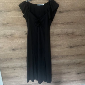 Svart långklänning från 157 - Svart långklänning från 157 med v-ringning och snygg knytning framtill. Klänningen har korta volangärmar och är tillverkad i ett lätt, krinklat tyg som ger en avslappnad känsla. Perfekt för dig som gillar stilrena och enkla plagg med en twist.