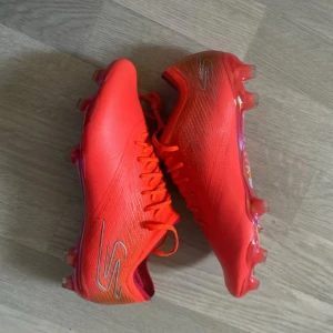 Skecher fotbollsskor - Snygga korallfärgade Skechers Razor fotbollsskor med snörning och cool, glansig sula. Skorna har en slimmad design och är tillverkade i syntetmaterial för bästa känsla på planen. Perfekta för dig som vill sticka ut med färg och stil under matchen.