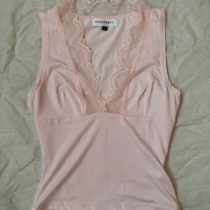 Ljusrosa linnetopp från Venderby’s - Supersöt ljusrosa linnetopp från Venderby’s med djup v-ringning och breda spetsdetaljer längs halsringningen. Toppen är ärmlös och har en figurnära passform i mjukt, stretchigt material. Aldrig använd, endast tvättad. Nypris ca 410kr