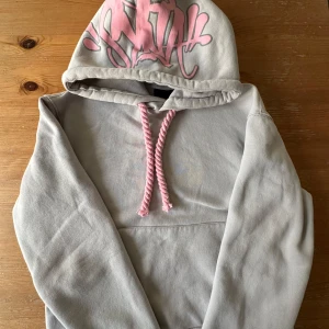 Grå hoodie med rosa tryck och snören - Säljer en grå hoodie från SYNA wrld med fet rosa graffiti-tryck på huvan och grova rosa snören. Tröjan har känguruficka framtill och är i mjuk bomullsmix. Perfekt för dig som gillar streetstyle och vill sticka ut med coola detaljer. 