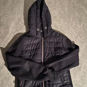 Svart quiltad jacka Tommy Hilfiger - Svart jacka från Tommy Hilfiger med quiltad framsida och huva. Jackan har dragkedja, ribbade muddar och små logodetaljer. Materialmix med vadderad front och mjuka ärmar. Perfekt för höst och vår när du vill ha en stilren look. Pris kan diskuteras vid snabb affär. 