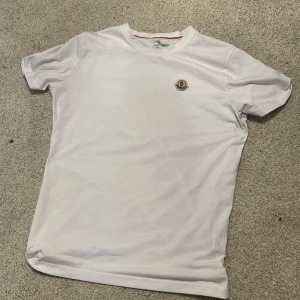 Vit Moncler t-shirt med logga - Vit t-shirt från Moncler i 100% bomull med klassisk rund hals och broderad logga på bröstet. Snygg detalj med röd, vit och blå rand på insidan av kragen. Enkel och clean design som passar till allt.