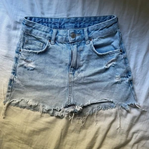 Ljusblå jeanskjol med slitningar - Säljer en kort ljusblå jeanskjol med fransig nederkant och slitna detaljer framtill. Kjolen har klassiska fickor och bälteshällor, perfekt för en avslappnad och trendig look.