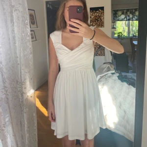 Vit omlottklänning  - Supersöt vit kortklänning från bubbelroom med omlottliv och breda axelband. Klänningen har en lätt utställd kjol och markerad midja, vilket ger en riktigt fin siluett. Perfekt för student eller skolavslutning Materialet är mjukt och följsamt. Storlek M