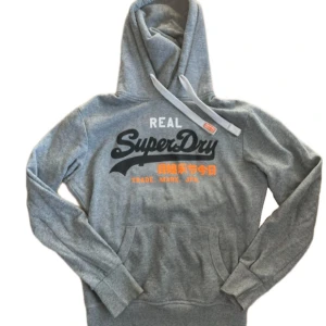 Grå hoodie från Superdry med tryck - Snygg grå hoodie från Superdry med stort svart och vitt logotryck samt orange japansk text på bröstet. Tröjan har huva med snörning och en klassisk känguruficka framtill. Mjuk insida i fleece för extra komfort.