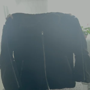 Svart teddyjacka med dragkedja - *Finns även i vit färg, storlek XL* Mysig svart teddyjacka med hög krage och hel dragkedja framtill. Jackan har två fickor på sidorna och är tillverkad i fluffigt teddy-material som håller dig varm. Perfekt för kyliga dagar och ger en cool, avslappnad vibe.