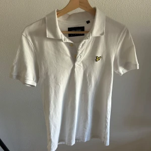 Vit piké från Lyle & Scott XS - Snygg vit piké från Lyle & Scott i storlek XS. Klassisk krage, korta ärmar och gul broderad logga på bröstet. Tröjan har knappar vid halsen och är gjord i mjuk bomull som känns skön mot huden.