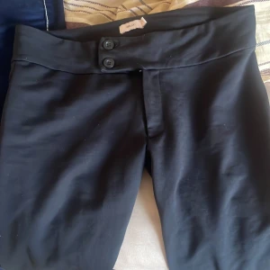 Svarta bootcut-byxor från Nelly - Snygga svarta bootcut-byxor från Nelly i storlek S. Byxorna har hög midja, två knappar framtill och är tillverkade i ett mjukt, stretchigt material som sitter skönt. Perfekt för dig som gillar en klassisk och stilren look. Nypris 350