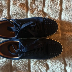 Mörkblå Christian Louboutin sneakers - Säljer ett par mörkblå sneakers från Christian Louboutin med klassisk röd sula och coola nitar på tån. Skorna har snörning och är gjorda i mocka med vita gummisulor. Perfekta för dig som vill sticka ut med exklusiv streetstyle.