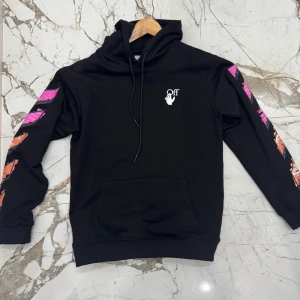 Svart Off-White hoodie med färgglada detaljer - Svart hoodie från Off-White med huva och snörning. Stora grafiska tryck i rosa och orange på ärmar och rygg, samt vit logga på bröstet. Känguruficka framtill och ribbade muddar. Tillverkad i mjuk bomull för maximal komfort.