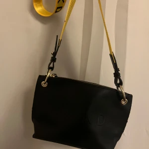 Svart axelväska från NA-KD med gul rem - Cool svart axelväska från NA-KD i skinnimitation med logga framtill. Väskan har en bred, justerbar axelrem i gult med svart text och snygga metalldetaljer i guld. Stängs med dragkedja upptill och har en modern, stilren look.