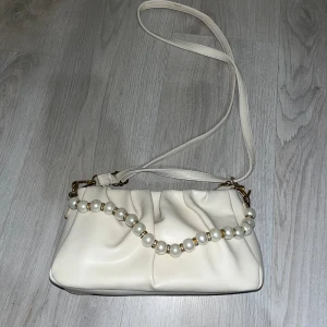 Vit axelväska med pärldetalj - Snygg vit axelväska i slätt syntetmaterial med veckad design och lång axelrem. Väskan har en dekorativ pärlkedja med gulddetaljer framtill som ger en lyxig känsla. Perfekt för dig som vill ha en trendig och stilren accessoar.