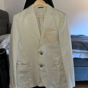 Beige kavaj i bomull - Stilren beige kavaj med två knappar framtill och tre knappar vid ärmslutet. Klassisk passform med två stora fickor och en bröstficka. Tillverkad i mjuk bomull, perfekt för en clean och avslappnad look. Däremot finns liten fläck vid bröstfickan, ska kunna fixas med kemtvätt. 