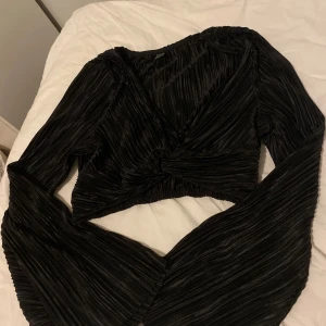 Svart omlottopp med vida ärmar - Säljer en svart omlottopp med plisserat tyg och vida långa ärmar. Toppen är croppad och har en snygg twist-detalj framtill. Perfekt för dig som vill sticka ut med en cool och trendig look.