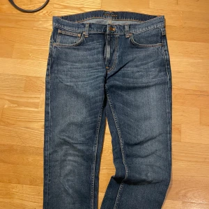 Blå jeans från Nudie Jeans, stl 32/30 - Säljer ett par klassiska blå jeans från Nudie Jeans i storlek 32/30. Jeansen har raka ben, fem fickor och snygga orangea sömmar samt Nudie-logga på bakfickan. Tillverkade i slitstarkt denim med normal passform och midja.