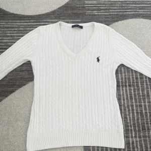 Vit kabelstickad tröja Polo Ralph Lauren - Vit kabelstickad tröja från Polo Ralph Lauren med v-ringning och klassisk marinblå logga broderad på bröstet. Tröjan har lång ärm och ribbade muddar, perfekt för en clean och stilren look.