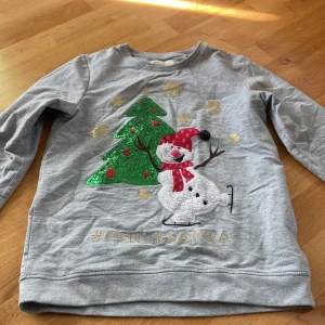 Grå jultröja Holly & Whyte Lindex - Grå långärmad tröja från Holly & Whyte by Lindex med broderad snögubbe, glittrig grön julgran och guldiga snöflingor. Texten #CHILLSANTA framtill. Perfekt för julens alla tillfällen.