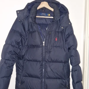 Mörkblå dunjacka Polo Ralph Lauren M - Mörkblå dunjacka från Polo Ralph Lauren med huva och dragkedja framtill. Jackan har broderad röd logga på bröstet, flera fickor med dragkedja och klassisk pufferstil. Perfekt för kalla dagar och riktigt snygg streetstil.