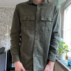 Olivgrön overshirt med fickor - Snygg olivgrön overshirt/jacka i bomull med klassisk krage och två bröstfickor med knappar. Perfekt inför höst/våren!