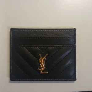 Svart YSL kortfodral i skinn - Supersnyggt svart kortfodral från YSL i äkta skinn med quiltad design och guldigt YSL-emblem framtill. Perfekt för att hålla koll på dina kort med stil. Slimmad och smidig att ha i fickan eller väskan.