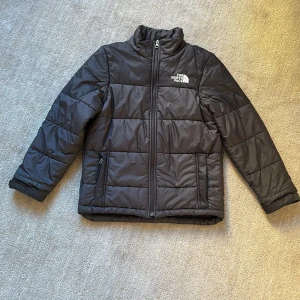 Svart  puffer jacka från The North face - Svart pufferjacka från The North Face (circulated synthetic jacket)  i storlek L (146-151) med polyesterfyllning. Jackan har hög krage, dragkedja framtill och två sidofickor med dragkedja. Klassisk quiltad design och logga på bröstet och ryggen. Perfekt för kyliga dagar och enkel att matcha med allt. Sparsamt använd, men det finns en pytteliten reva på 1,5 cm framtill. Enkelt att fixa. 