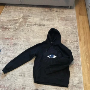 Svart Kenzo hoodie med ögonmotiv - Svart hoodie från Kenzo med ikoniskt broderat öga på bröstet och texten 'Paris' i motivet. Tröjan har huva med snörning och klassisk känguruficka. Perfekt för dig som gillar streetwear och vill sticka ut med en unik detalj. Oanvänd helt ny. Passar även i S