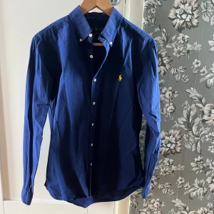 Blå skjorta från Ralph Lauren M - Klassisk marinblå skjorta från Ralph Lauren i slim fit-modell. Skjortan har långa ärmar, knappar framtill och den ikoniska gula loggan broderad på bröstet. Tillverkad i bomull och passar perfekt för en stilren look.