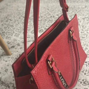 Röd handväska i skinnimitation - Snygg röd handväska med struktur som påminner om ormskinn. Väskan har både handtag och axelrem, guldfärgade metalldetaljer och dragkedja upptill. Perfekt för dig som vill sticka ut med en färgstark accessoar. Dekorativ tag med texten 'LOVE' hänger på handtaget.