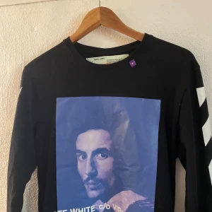 Svart långärmad Off-White tröja - Svart långärmad tröja från Off-White med vit text och grafiskt tryck i blått framtill. Klassiska vita pilar på ärmarna och tryck på ryggen med 'BUSINESS CASUAL'. Crewneck och relaxed fit, perfekt för dig som gillar streetwear.