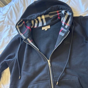Svart Burberry hoodie med dragkedja - Svart hoodie från Burberry i storlek M med klassiskt rutigt foder i huvan och snygga metalldetaljer på snöret. Burrbery tröjan är äkta och kvitto finns bara kontakta mig, samt pris kan diskuteras i fall det är ett bra pris förslag.