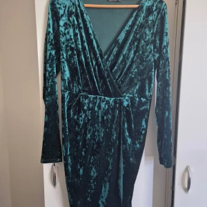 Mörkgrön sammetsklänning NLY One XL - Supersnygg mörkgrön sammetsklänning från NLY One i omlottmodell. Klänningen har lång ärm, v-ringning och markerad midja med knytband. Materialet är glansigt och mjukt, vilket ger en lyxig känsla. Perfekt för dig som vill sticka ut med en elegant look.